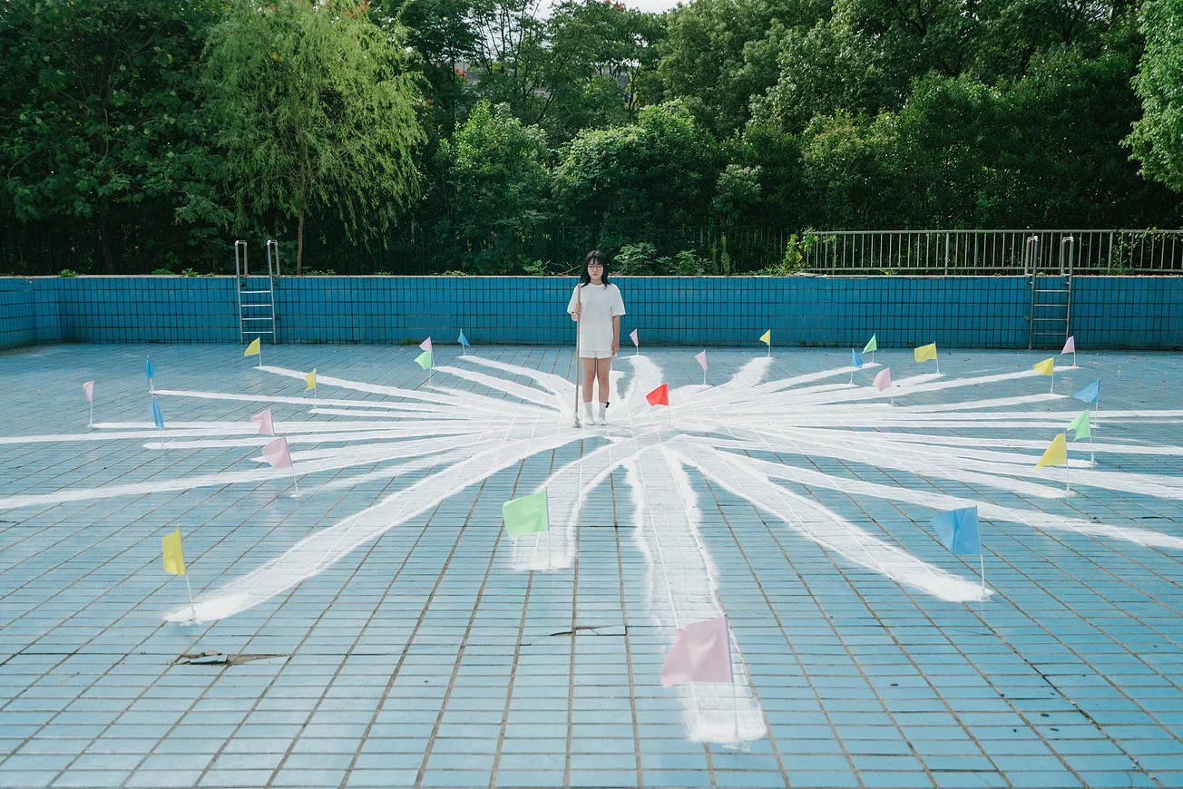 A woman stands in the centre of an empty swimming pool where colourful flags are placed around the space. She has drawn lines from the centre to each flag with a white powder, creating a star pattern on the floor.